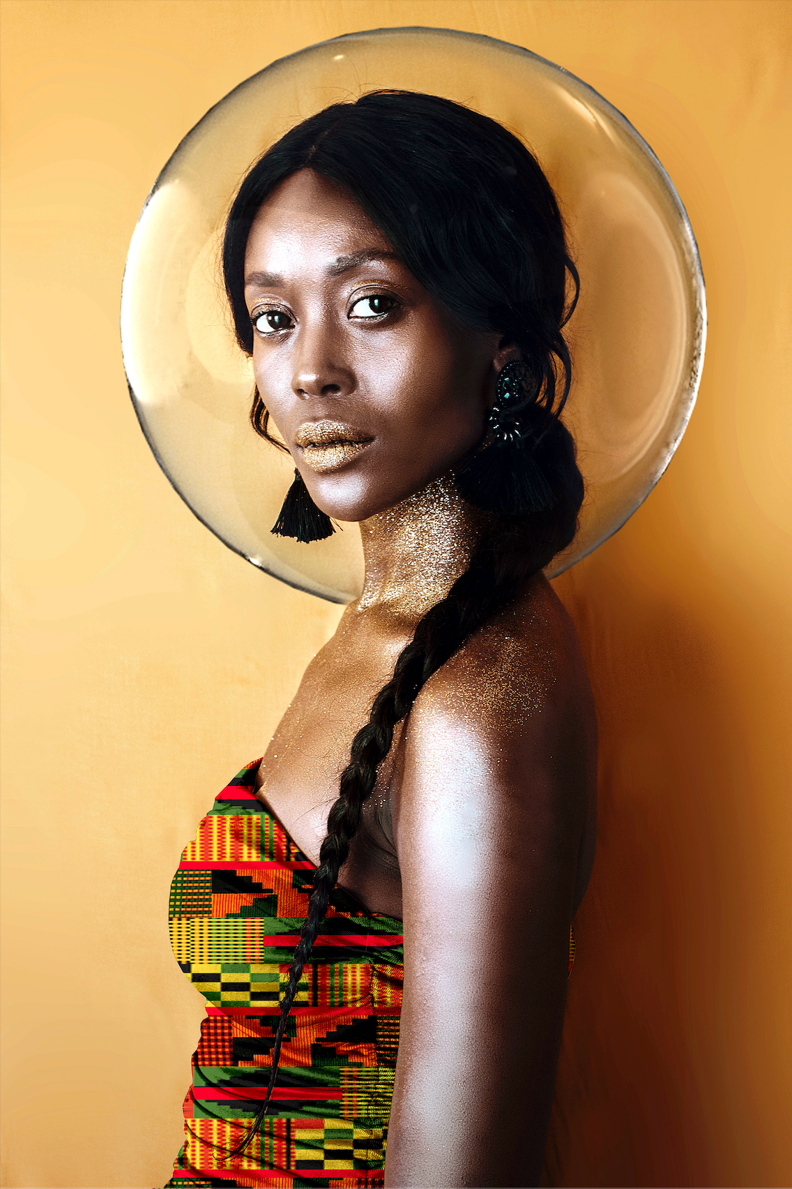 Woman wearing a colorful African Kente dress with a reflective circular object in front of her face on a orange background